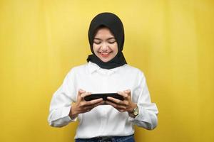 Beautiful asian young muslim woman with hands holding smartphone, playing game, smiling happily, victory, success, isolated on yellow background, advertising concept photo