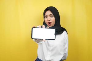 Beautiful young asian muslim woman shocked, surprised, wow expression, hand holding smartphone with white or blank screen, promoting app, promoting product, presenting something, isolated photo