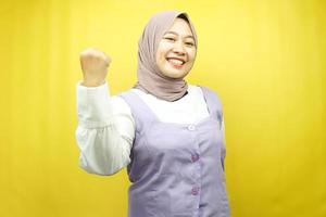 Beautiful young asian muslim woman smiling confident, enthusiastic and cheerful with hands clenched, sign of success, punching, fighting, not afraid, victory, isolated on yellow background photo