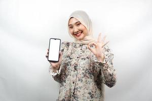 Hermosa joven musulmana asiática sonriendo con confianza y entusiasmo con las manos sosteniendo el teléfono inteligente, promoviendo la aplicación, mano de signo ok, buen trabajo, éxito, aislado sobre fondo blanco. foto