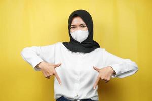 Muslim woman smiling confidently pointing down, presenting something, click the link below, isolated on yellow background photo
