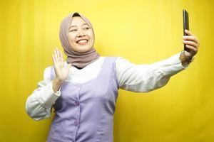 Hermosa joven musulmana asiática sonriendo confiada, entusiasta y alegre con las manos sosteniendo el teléfono inteligente, llamando a alguien, haciendo videollamadas, tomando selfie, aislado sobre fondo amarillo foto