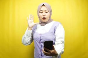 Hermosa joven musulmana asiática conmocionada, sorprendida, con las manos sosteniendo el teléfono inteligente, mirando el teléfono inteligente, mirando la promoción, aislado sobre fondo amarillo, concepto de publicidad foto