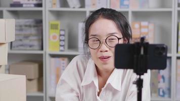 Happy young Asian woman wearing glasses and sitting to do live on social media at home, setting camera on a table, Women doing business selling online, online sales channel, woking at home video