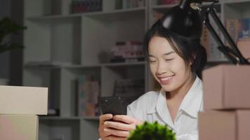 Feliz joven asiática recibiendo mensajes del cliente en el teléfono inteligente, teniendo nuevos pedidos en línea, niña sentada en la sala de trabajo por la noche. trabajando en casa. venta de concepto en línea video