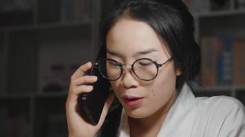 Close up face of Asian woman wearing glasses taking a call about work in working room at night, explaining more details, working hard, working from home concept
