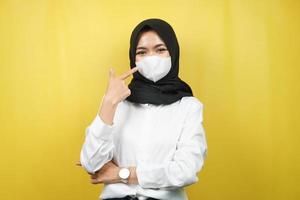 Mujer musulmana con máscara blanca, con la mano apuntando a la máscara, prevenir el gesto del virus corona, prevenir el covid-19, aislado sobre fondo amarillo