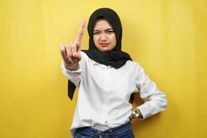 Beautiful assertive asian young muslim woman, with number one sign hand, warning sign hand to camera, isolated on yellow background