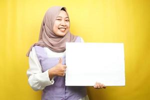 Bastante joven mujer musulmana alegre sosteniendo pancarta vacía en blanco, cartel, tablero blanco, tablero de letrero en blanco, tablero de anuncio blanco, presentando algo en el espacio de la copia, promoción foto