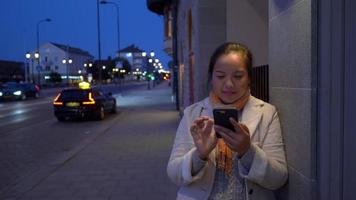 Happy Asian woman standing and using smartphone on the footpath in the city at night. Slide on the screen and texing on phone on the street. Building and moving car background. City night concept video