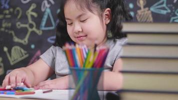 Vista frontal de la feliz niña asiática sentada y pintando con lápiz de color sobre la mesa. haciendo actividades artísticas el fin de semana solo en la sala de estudio de casa.