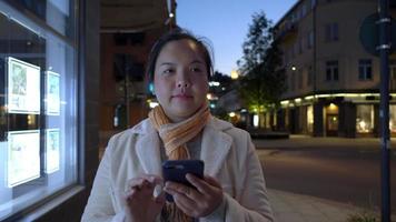 aziatische vrouw die sjaal draagt en smartphone op het voetpad in de stad gebruikt. schuif op het scherm en sms op de telefoon op straat. gebouw en bewegende auto achtergrond. stad nacht concept video