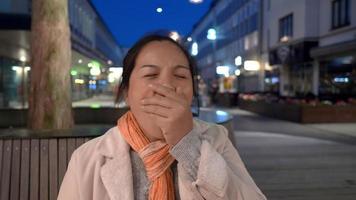 vista frontale della donna asiatica in piedi e in attesa di un amico, chiudendo la bocca e sbadigliando. stare da solo in città di notte. edificio e sfondo chiaro di notte. concetto di notte della città video