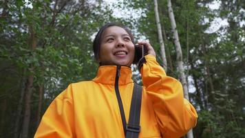 Happy Asian black hair woman wearing yellow coat taking a call in the forest with big green trees, traveling outdoor in beautiful nature. Big trees background. Forest concept