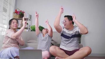 feliz familia asiática sentada en el suelo en la sala de estar. jugando y bailando juntos, poniendo la computadora portátil frente a ellos. hija, mamá y papá riendo y pasando un buen rato. concepto de familia feliz video