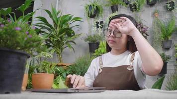 Une vendeuse de plantes asiatique finit de travailler, ferme l'ordinateur portable, enlève les lunettes et met sur la table puis se repose. travailler comme vendeur d'arbres à la maison, se fatiguer video
