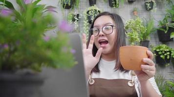 Feliz joven asiática vendiendo plantas en línea, sosteniendo productos vegetales y usando una computadora portátil para explicar los detalles a su cliente en vivo en las redes sociales. venta de concepto en línea. video