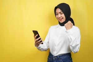 Hermosa joven musulmana asiática sonriendo confiada, entusiasta y alegre con las manos sosteniendo el teléfono inteligente, apretando las manos, puñetazos, victoria, éxito, buen trabajo, aislado sobre fondo amarillo