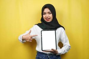 Beautiful young asian muslim woman smiling, excited and cheerful holding tablet with white or blank screen, promoting app, promoting product, presenting something, isolated on yellow background photo