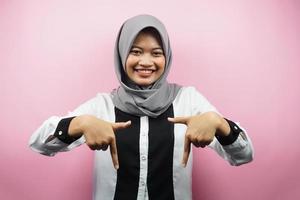 Beautiful asian young muslim woman with hands pointing down, hands click the link below, hands presenting something, isolated on pink background photo