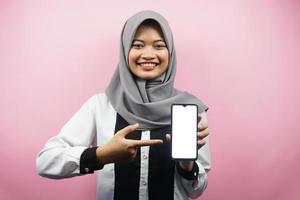 Beautiful young asian muslim woman smiling confident, enthusiastic and cheerful with hand pointing at smartphone, presenting something, hand presenting app, isolated on pink background