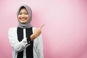 Beautiful young asian muslim woman with hands pointing empty space presenting something, smiling confident, enthusiastic, cheerful, looking at camera, isolated on pink background, advertising concept photo