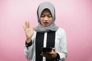 Beautiful young asian muslim woman shocked, surprised, with hands holding smartphone, looking at smartphone, looking at promo, isolated on pink background, advertising concept photo