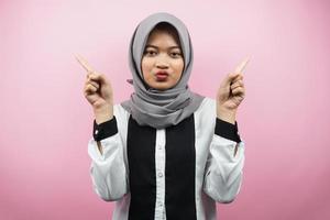 Beautiful asian young muslim woman with mouth watering, hand pointing at empty space presenting something, isolated on pink background photo