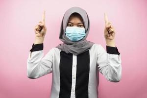 Muslim woman wearing medical mask, hand pointing to empty space, hand pointing up presenting something, isolated on pink background photo