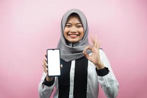 Beautiful young asian muslim woman smiling confidently and excitedly with hands holding smartphone, presenting application, ok sign hand, good job, success, isolated on pink background photo