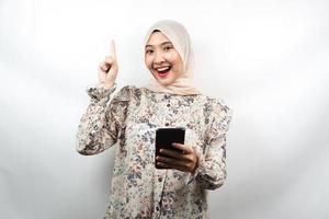Hermosa joven mujer musulmana asiática sonriendo, conmocionada, sorprendida, teniendo una idea, con las manos sosteniendo el teléfono inteligente, aislado sobre fondo blanco, concepto de publicidad foto