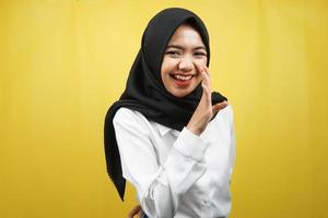 Beautiful young asian muslim woman smiling confidently and excitedly close to camera, whispering, telling secrets, speaking quietly, silent, isolated on yellow background photo