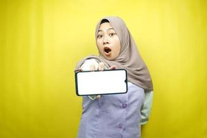 Hermosa joven mujer musulmana asiática conmocionada, sorprendida, expresión guau, mano sosteniendo un teléfono inteligente con pantalla blanca o en blanco, promocionando la aplicación, promocionando el producto, presentando algo, aislado foto
