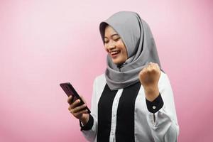 Hermosa joven musulmana asiática sonriendo confiada, entusiasta y alegre con las manos sosteniendo el teléfono inteligente, apretando las manos, puñetazos, victoria, éxito, buen trabajo, aislado sobre fondo rosa foto