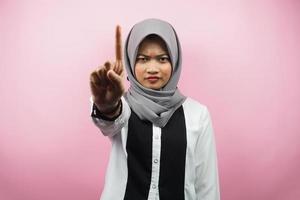 Beautiful assertive asian young muslim woman, with number one sign hand, warning sign hand to camera, isolated on pink background