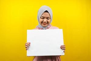 Bastante joven mujer musulmana alegre sosteniendo pancarta vacía en blanco, cartel, tablero blanco, tablero de letrero en blanco, tablero de anuncio blanco, presentando algo en el espacio de la copia, promoción foto