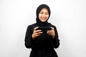 Beautiful asian young muslim woman with hands holding smartphone, playing game, smiling happily, victory, success, isolated on white background, advertising concept photo