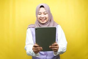 Hermosa joven mujer musulmana asiática sonriente, emocionada y alegre sosteniendo la tableta, aislado sobre fondo amarillo foto