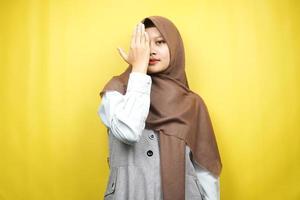 Beautiful asian young muslim woman with hand covering one eye looking at camera isolated on yellow background photo