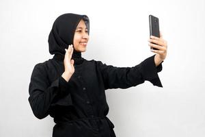 Hermosa joven musulmana asiática con manos sosteniendo un teléfono inteligente, llamando a alguien, haciendo videollamadas, tomando selfie, sonriendo confiado, entusiasta y alegre aislado sobre fondo blanco. foto