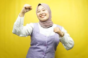 Beautiful young asian muslim woman smiling confident, enthusiastic and cheerful with  hands clenched, sign of success, punching, fighting, not afraid, isolated on yellow background photo