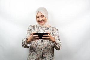 Beautiful asian young muslim woman with hands holding smartphone, playing game, smiling happily, victory, success, isolated on white background, advertising concept photo