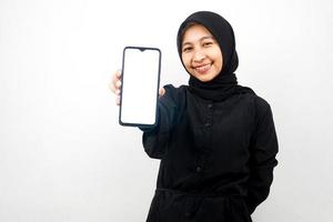 Hermosa joven musulmana asiática con las manos sosteniendo el teléfono inteligente, promoviendo la aplicación, promocionando algo, sonriendo confiada, entusiasta y alegre, aislado sobre fondo blanco.