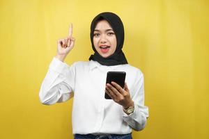 Beautiful young asian muslim woman smiling, shocked, surprised, getting an idea, with hands holding smartphone, isolated on yellow background, advertising concept