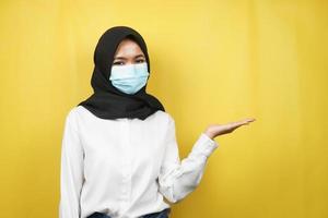 Mujer musulmana con máscara médica, manos presentando algo en el espacio vacío, aislado sobre fondo amarillo