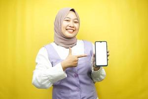 Hermosa joven musulmana asiática sonriendo confiada, entusiasta y alegre con la mano apuntando al teléfono inteligente, promocionando algo, aplicación de promoción manual, aislada sobre fondo amarillo foto