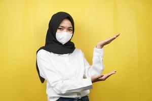 Mujer musulmana con máscara blanca, con la mano apuntando al espacio vacío presentando algo, aislado sobre fondo amarillo