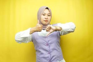 Beautiful asian young muslim woman with hand sign like or dislike, yes or no, happy or sad, comparing two things, isolated on yellow background photo