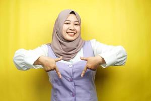 Beautiful asian young muslim woman with hands pointing down, hands click the link below, hands presenting something, isolated on yellow background photo