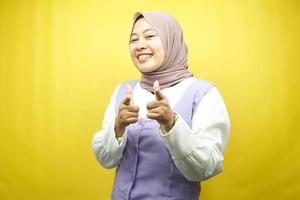 Beautiful young asian muslim woman smiling confident, enthusiastic and cheerful with hands pointing at camera, hands pointing at audience, facing camera isolated on yellow background photo